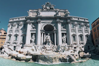 An ornate and grand baroque-style fountain adorned with intricate sculptures and statues, set against a backdrop of a historic building with majestic columns and decorative architecture.