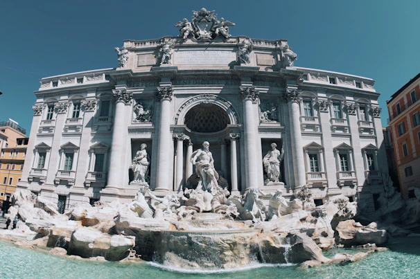 An ornate and grand baroque-style fountain adorned with intricate sculptures and statues, set against a backdrop of a historic building with majestic columns and decorative architecture.