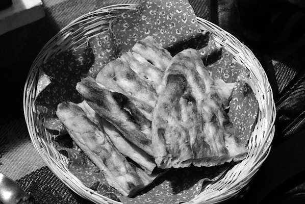 A woven basket contains several pieces of sliced flatbread. The flatbread appears toasted and is placed on top of a patterned paper or fabric lining the basket. The texture of the bread looks crisp and uneven, suggesting a freshly baked appearance.