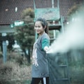 A young woman is standing beside a tennis net, holding it with one hand. She is smiling and looking at the camera. She wears a casual outfit with a graphic t-shirt and leggings. In the background, a partially visible building and some foliage are present, with a structure resembling a ladder or umpire chair.