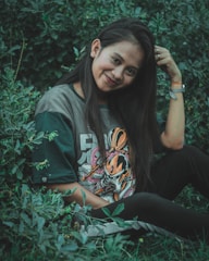 A young woman with long dark hair is sitting in lush green foliage. She is wearing a t-shirt with a colorful graphic design and smiling at the camera, gently touching her hair. Her relaxed pose and the natural surroundings contribute to a serene atmosphere.