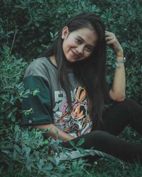 A young woman with long dark hair is sitting in lush green foliage. She is wearing a t-shirt with a colorful graphic design and smiling at the camera, gently touching her hair. Her relaxed pose and the natural surroundings contribute to a serene atmosphere.