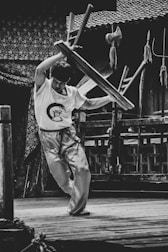 A person, dressed in martial arts attire, is energetically lifting a stool above their head in an outdoor setting. The background features traditional wooden architecture and various martial arts weapons are displayed nearby.