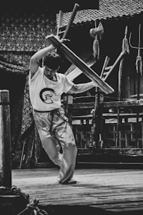 A person, dressed in martial arts attire, is energetically lifting a stool above their head in an outdoor setting. The background features traditional wooden architecture and various martial arts weapons are displayed nearby.