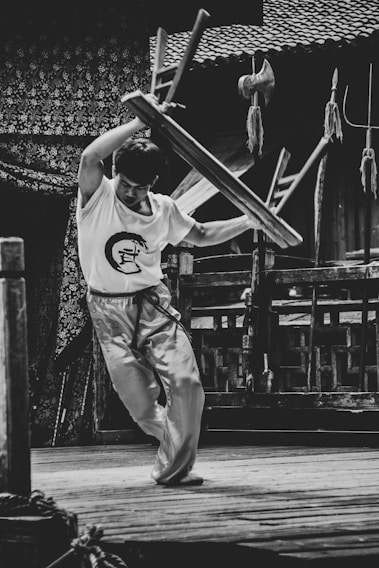 A person, dressed in martial arts attire, is energetically lifting a stool above their head in an outdoor setting. The background features traditional wooden architecture and various martial arts weapons are displayed nearby.