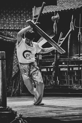 A person, dressed in martial arts attire, is energetically lifting a stool above their head in an outdoor setting. The background features traditional wooden architecture and various martial arts weapons are displayed nearby.