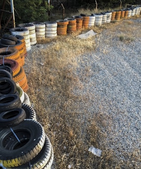 A long line of stacked tires in various colors, including black, white, and orange, wraps around a gravel and dry grass area. The tires are arranged neatly in layers, possibly for storage or use as a barrier.