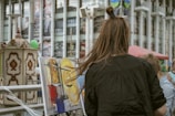 A person with long brown hair is painting on a canvas set up on a street. The painting features abstract swirls of yellow, blue, and red. In the background, there are columns and a carousel-like structure, along with advertisements.
