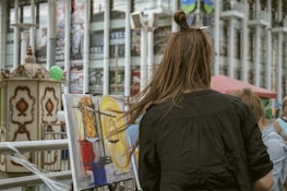 A person with long brown hair is painting on a canvas set up on a street. The painting features abstract swirls of yellow, blue, and red. In the background, there are columns and a carousel-like structure, along with advertisements.