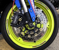 A close-up view of a vibrant neon yellow motorcycle wheel featuring a dual-disc brake system. The brake caliper is prominently marked with the brand 'Tokico' and is mounted on a blue suspension fork. The wheel's spokes are visible through the large disc rotors, which have numerous holes for improved heat dissipation.