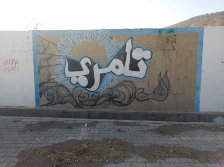 A section of a wall features a piece of street art with Arabic calligraphy in large, bold letters. The artwork is framed by intricate designs and decorative patterns, with a background in shades of beige and blue. Additional text appears to be graffiti on the left side of the wall.