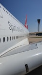 A close-up view of the side of a passenger airplane with the words 'Spirit of Australia' visible. The aircraft's body is white with a series of windows running along its side. In the background, there is an airport control tower under a clear blue sky.