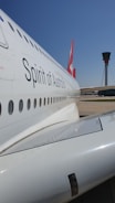 A close-up view of the side of a passenger airplane with the words 'Spirit of Australia' visible. The aircraft's body is white with a series of windows running along its side. In the background, there is an airport control tower under a clear blue sky.