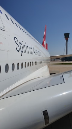 A close-up view of the side of a passenger airplane with the words 'Spirit of Australia' visible. The aircraft's body is white with a series of windows running along its side. In the background, there is an airport control tower under a clear blue sky.