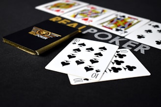 A deck of playing cards labeled 'Real Poker' rests on a dark surface. Two cards, the ten of spades and ten of clubs, are prominently displayed in the foreground. A row of face cards, including kings and queens from different suits, is lined up in the background.