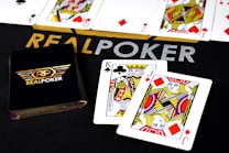 Poker-themed setup featuring a deck of cards on a table with the words 'REAL POKER' visible. Two cards, the King of Clubs and Jack of Diamonds, are prominently displayed. The design is vibrant, with detailed illustrations on the cards.