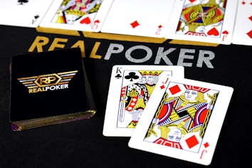 Poker-themed setup featuring a deck of cards on a table with the words 'REAL POKER' visible. Two cards, the King of Clubs and Jack of Diamonds, are prominently displayed. The design is vibrant, with detailed illustrations on the cards.
