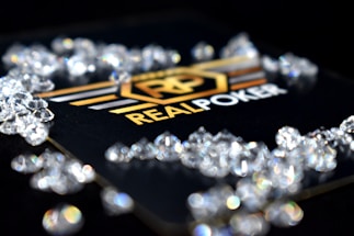 A collection of sparkling, clear gemstones surrounds a dark surface featuring the prominent logo and text 'Real Poker.' The logo design includes gold and white elements set against a black background.