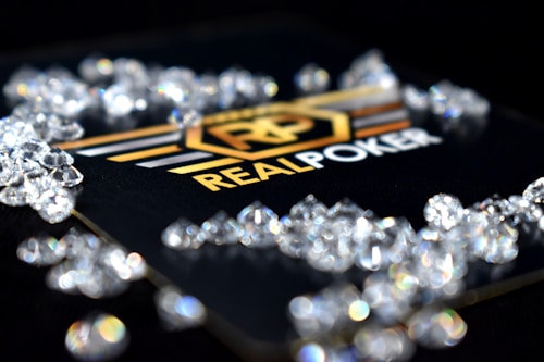 A collection of sparkling, clear gemstones surrounds a dark surface featuring the prominent logo and text 'Real Poker.' The logo design includes gold and white elements set against a black background.