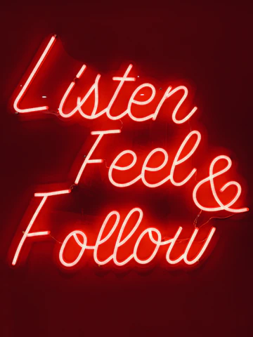 Bright red neon sign displaying the words 'Listen Feel & Follow' in cursive script on a dark background.
