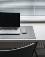 A minimalist workspace featuring a closed laptop and a smartphone resting on a dark desk mat. The items are placed on a sleek, white desk positioned in front of a window with closed blinds. The scene conveys a sense of simplicity and modernity.