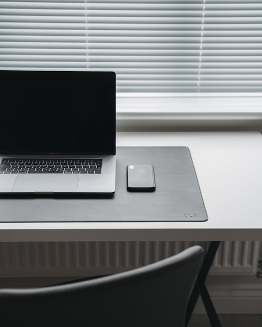 A minimalist workspace featuring a closed laptop and a smartphone resting on a dark desk mat. The items are placed on a sleek, white desk positioned in front of a window with closed blinds. The scene conveys a sense of simplicity and modernity.