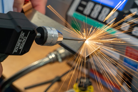 Sparks fly from a piece of metal being worked on with a power tool, suggesting a metalworking or welding process in a workshop setting. A caution label indicates the tool produces heat.