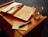 An open book with ribbon bookmarks lies on a wooden surface next to a transparent mug containing a small amount of liquid. A handwritten note on a piece of paper and a blue pen accompany the book.