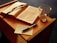 An open book with ribbon bookmarks lies on a wooden surface next to a transparent mug containing a small amount of liquid. A handwritten note on a piece of paper and a blue pen accompany the book.