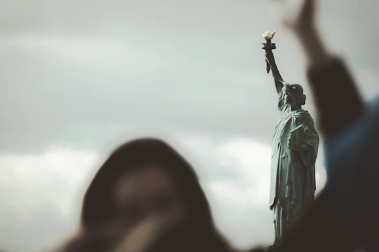 A warm, inviting photo of the Statue of Liberty with Ellis Island softly blurred in the background at sunset.