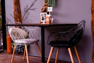 A cozy dining setup featuring two modern, wire-frame chairs, one white and one black, placed around a dark wooden table. The table is set against a gray wall with natural wooden beams and some decorative greenery. A table marker labeled 'C4', a box of tissues, and condiments are placed on the table. An orange gas cylinder can be seen at the side.