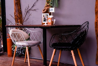 A cozy dining setup featuring two modern, wire-frame chairs, one white and one black, placed around a dark wooden table. The table is set against a gray wall with natural wooden beams and some decorative greenery. A table marker labeled 'C4', a box of tissues, and condiments are placed on the table. An orange gas cylinder can be seen at the side.