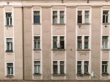 A beige residential building facade with multiple windows aligned in columns. Most of the windows have white curtains, while a few are open or partially open. Some windows feature flower boxes with greenery.