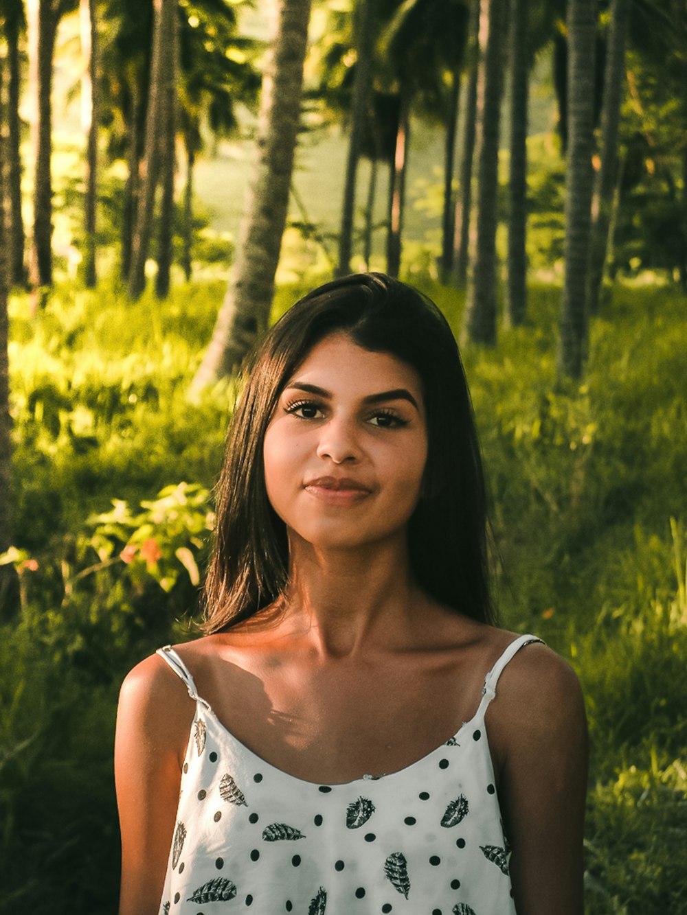 woman wearing white and black spaghetti strap top standing on green field surrounded with tall and green trees