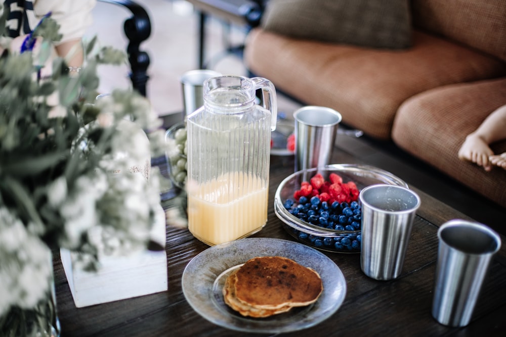 four gray cups beside bowls with berries and pitcher with half-empty juice near pancakes on table