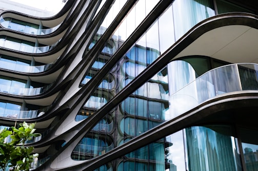 A modern building with a futuristic design featuring curved glass facades and metallic elements. The structure exhibits a combination of sleek lines and reflective surfaces, creating an architectural flow. Trees with green leaves are visible in the foreground, adding a touch of nature to the urban scene.
