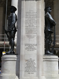 A war memorial features two statues of soldiers in uniform, standing on either side of an inscribed stone pillar. The inscription honors the officers and men of London who served in the Great War, and it mentions unveiling dates and commemorations. The architecture is grand, with columns visible in the background.