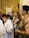 A group of people are gathered indoors, appearing attentive or engaged in an event or exhibition. Some are holding brochures or pamphlets, and they seem to be standing in a hall with art pieces displayed in the background.