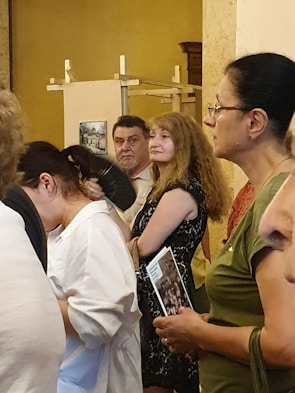 A group of people are gathered indoors, appearing attentive or engaged in an event or exhibition. Some are holding brochures or pamphlets, and they seem to be standing in a hall with art pieces displayed in the background.