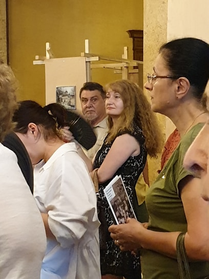 A group of people are gathered indoors, appearing attentive or engaged in an event or exhibition. Some are holding brochures or pamphlets, and they seem to be standing in a hall with art pieces displayed in the background.