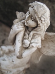 A weathered, stone angel sculpture with delicate features and wings folded behind. The angel appears thoughtful, holding a small object close to its chest. The surface is textured with signs of age and dirt, adding a rustic charm.