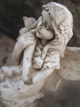 A weathered, stone angel sculpture with delicate features and wings folded behind. The angel appears thoughtful, holding a small object close to its chest. The surface is textured with signs of age and dirt, adding a rustic charm.