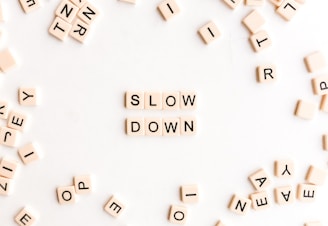 A collection of letter tiles scattered on a white surface with the words 'SLOW DOWN' formed in the center. The tiles are cream-colored with black letters, creating a contrast against the backdrop.