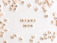 A collection of cream-colored Scrabble-like tiles with black letters scattered on a white surface. At the center, tiles are arranged to form the words 'START NOW'.