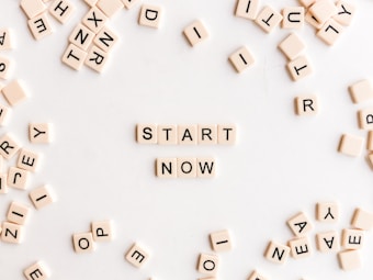 A collection of cream-colored Scrabble-like tiles with black letters scattered on a white surface. At the center, tiles are arranged to form the words 'START NOW'.