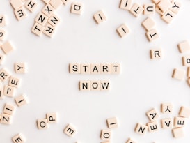 A collection of cream-colored Scrabble-like tiles with black letters scattered on a white surface. At the center, tiles are arranged to form the words 'START NOW'.