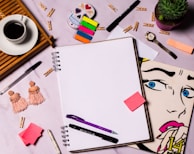A flat lay of a creative workspace featuring a blank spiral notebook surrounded by various items including colorful sticky notes, pens, a ceramic cup of coffee on a wooden tray, a pair of tasseled earrings, a succulent plant in a purple pot, a wristwatch, a stylized pop art drawing of a woman's face, and a decorative skull. Small wooden clothespins are scattered across the surface.