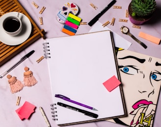 A flat lay of a creative workspace featuring a blank spiral notebook surrounded by various items including colorful sticky notes, pens, a ceramic cup of coffee on a wooden tray, a pair of tasseled earrings, a succulent plant in a purple pot, a wristwatch, a stylized pop art drawing of a woman's face, and a decorative skull. Small wooden clothespins are scattered across the surface.