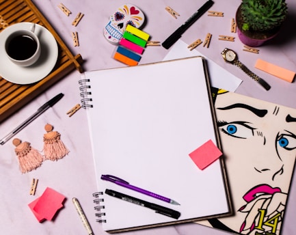 A flat lay of a creative workspace featuring a blank spiral notebook surrounded by various items including colorful sticky notes, pens, a ceramic cup of coffee on a wooden tray, a pair of tasseled earrings, a succulent plant in a purple pot, a wristwatch, a stylized pop art drawing of a woman's face, and a decorative skull. Small wooden clothespins are scattered across the surface.
