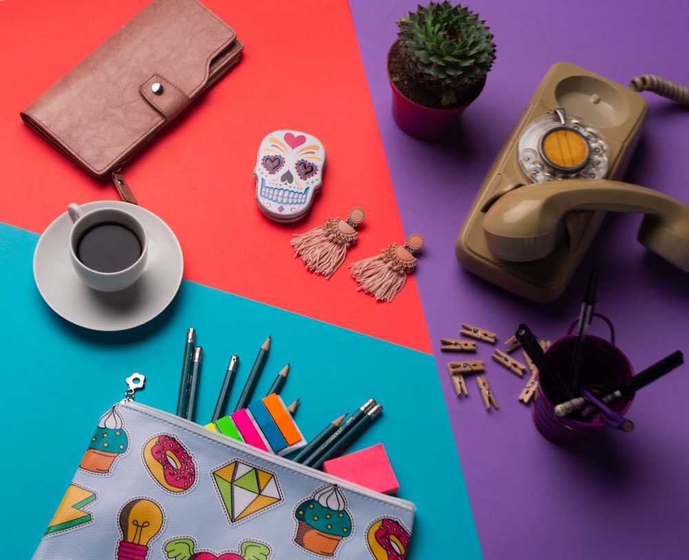 pencils on pouch beside coffee cup with saucer, cup with ballpoin pens near brown telephone, succulent plants, kalavera case, and brown wallet
